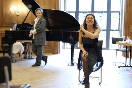 Frau auf einem Stuhl schaut in die Kamera, im Hintergrund ein Flügel