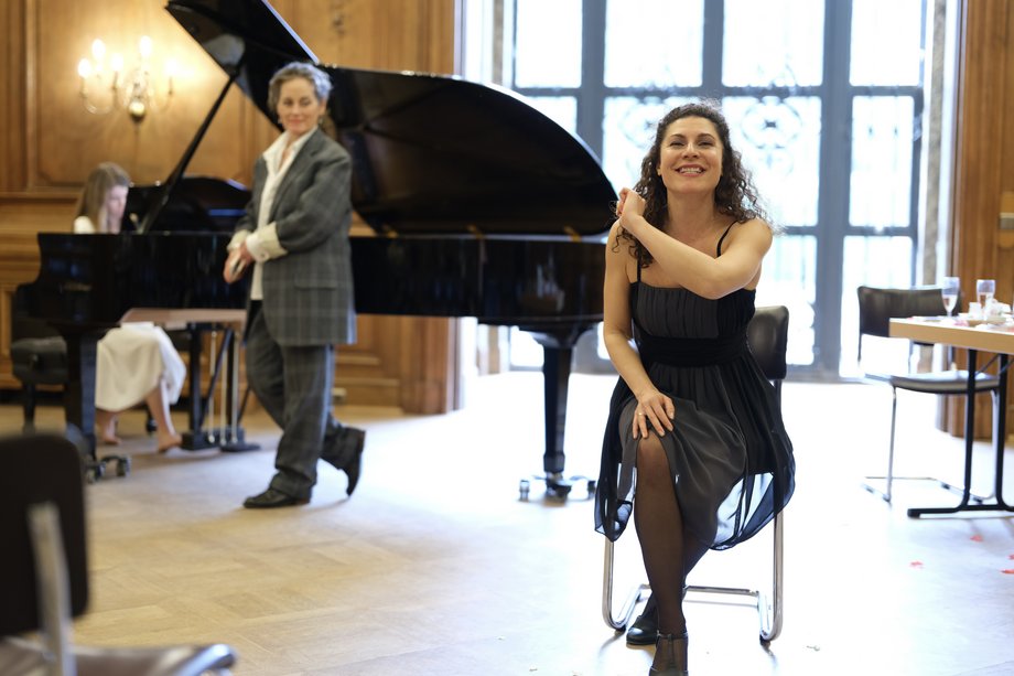 Frau auf einem Stuhl schaut in die Kamera, im Hintergrund ein Flügel