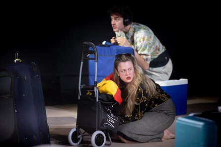 Anouk Piwek beim Schauspielschultreffen 2025 in Salzburg Eine Frau kniet vor einem Einkaufstrolley, hinter ihr ein Mann mit blauem Koffer
