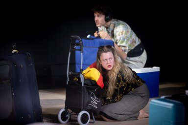 Anouk Piwek beim Schauspielschultreffen 2025 in Salzburg A woman kneels in front of a shopping trolley, behind her a man with a blue suitcase