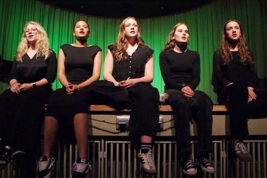 2025-11-02JungeKonzerte 5 Jugendliche sitzen auf dem Rand eine Bühne und singen vor grünem Hintergrund