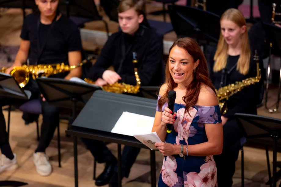 2025-11-18JugendJazzt Frau mit Mikrofon, im Hintergrund Jugendliche mit Instrumenten