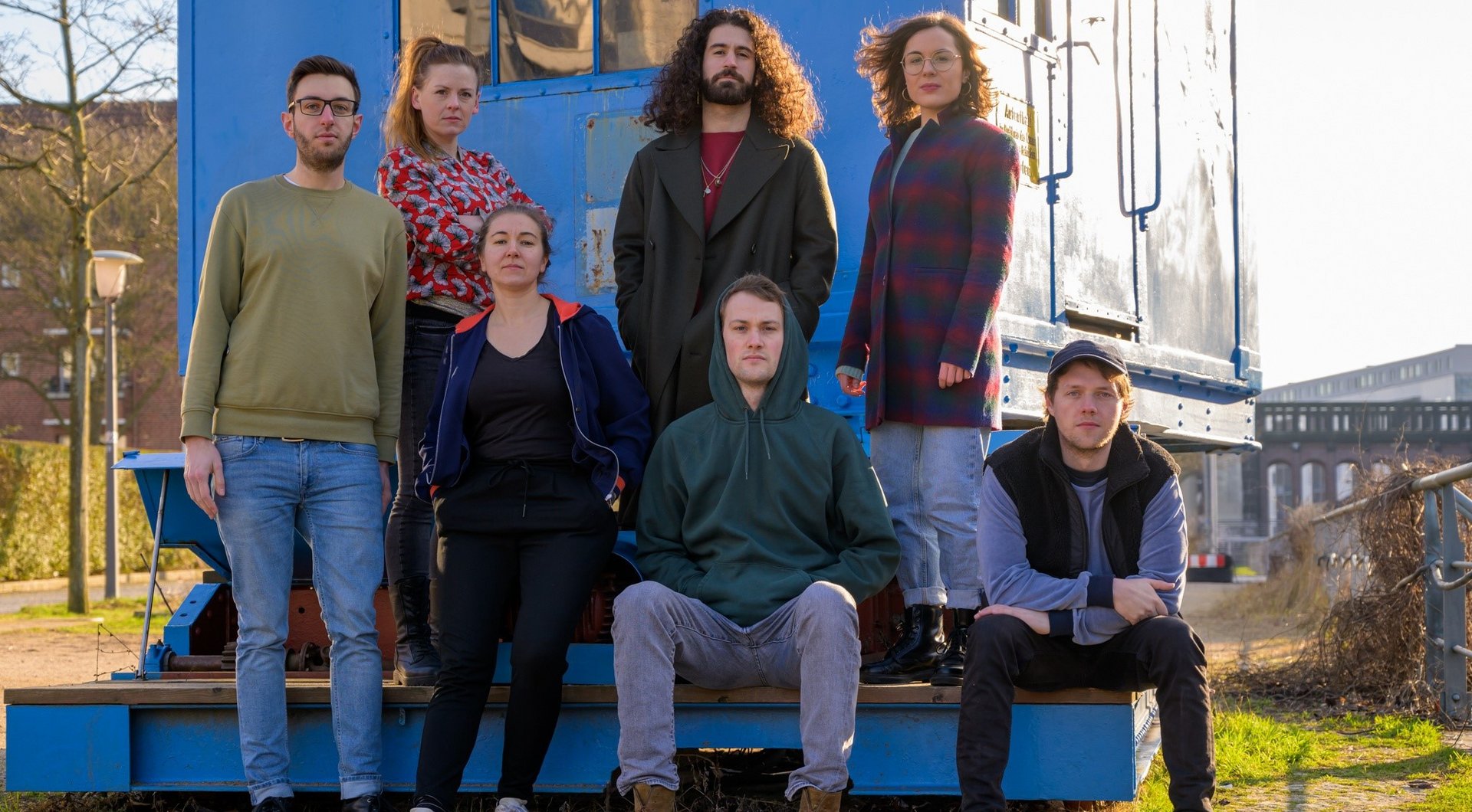 7 junge Menschen vor blauem Baucontainer, strahlendes Wetter