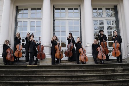 Das Ensemble mit den Celli auf einer Treppe stehend zeigt mit Fingern zur Kamera