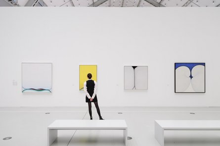 Huguette Caland 3 zu 2 Frau von hinten in einem großen Ausstellungshalle vor 3 Bildern