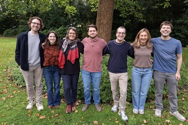 Seven people are standing side by side in a meadow, hugging each other