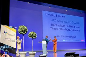 Abschlussveranstaltung AEC Salzburg Drei Personen stehen auf einer Bühne und im Hintergrund ist eine Leinwand