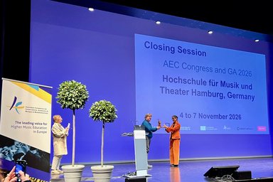 Drei Personen stehen auf einer Bühne und im Hintergrund ist eine Leinwand